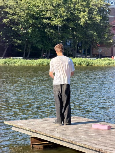 A person stands on a wooden dock, facing away from the camera, looking out at a body of water. The person is wearing a white t-shirt and dark pants. The water is rippling, reflecting the sunlight. Behind the dock, there is a line of tall grass and then trees. In the background, a building with signage is visible. The scene appears to be set during the day, with bright sunlight casting shadows. The overall mood is peaceful and still. There is a small pink rectangular object on the dock near the person. A thin white line extends from the person and disappears into the water.