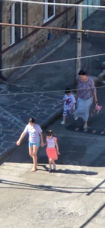This media file depicts a group of people in what appears to be an outdoor setting with a stone-paved ground and some industrial or structural elements in the background. Four individuals are visible, oriented vertically from bottom to top.

The bottommost subject is a person wearing a light-colored t-shirt with a graphic and blue shorts, with their arms extended. Above them is a person in a pink skirt and a light-colored top, also with arms outstretched. Next is a child in a white t-shirt with a red and blue graphic. The topmost visible person is an adult wearing a purple patterned blouse and a light blue skirt, holding a pink object in their hand. All individuals appear to be walking or posing.

The scene is lit by sunlight, casting distinct shadows on the ground, suggesting it is daytime. The presence of industrial structures and the stone paving might indicate a public area, a promenade, or an area near a port or construction site. The exact time of day and weather conditions are not explicitly discernible beyond the presence of daylight. No visible text is present in the image. The overall mood is neutral, with the individuals seemingly engaged in everyday activity.