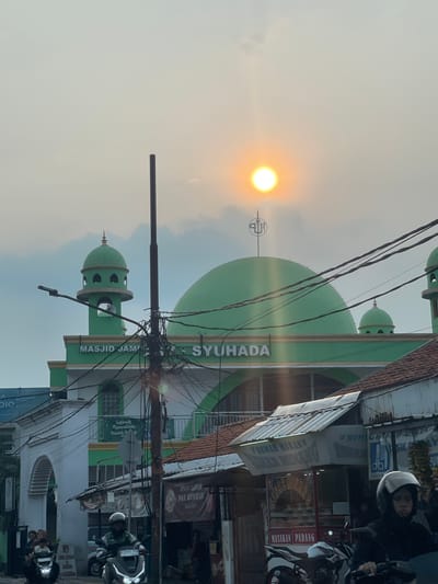 The image depicts the "Masjid Jami' Syuhada" mosque in South Tangerang, Indonesia, during what appears to be sunset. The mosque features a prominent green dome and a minaret, both characteristic of Islamic architecture. The sun, a bright orb with visible rays, is setting in the hazy sky, casting a warm glow on the scene. In the foreground, a street scene unfolds with several motorcycles, some with riders, navigating the road. Overhead, a tangle of electrical wires crisscrosses the frame, a common sight in urban environments. Buildings line the street, including what appears to be small businesses or shops. The overall atmosphere is that of a bustling urban environment with a prominent religious structure, set against the backdrop of a dramatic sunset.