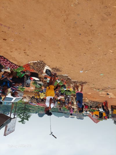 This is an outdoor scene of a busy market in Awkuzu, Nigeria, with a dirt ground. The image is oriented sideways. In the foreground and middle ground, there are numerous people, predominantly women, engaged in selling and buying goods. Stalls are set up with various produce, including piles of red onions, tomatoes, and what appear to be yams or similar root vegetables. Some women are seated, while others are standing and interacting with customers or fellow vendors. There are also vehicles visible, including a white van and a yellow taxi. The background shows a hazy sky and a treeline with buildings in the distance, suggesting an urban or semi-urban environment. The overall atmosphere is one of a bustling local marketplace.