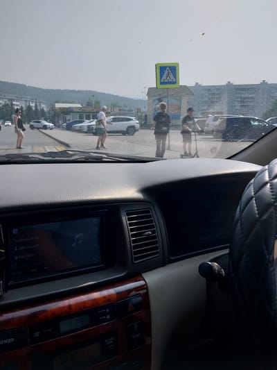 The image shows a view from inside a car, looking out at a street scene. A pedestrian crossing is visible, with several people crossing. One person is on a scooter. There are buildings in the background, and it appears to be a sunny day. The car's dashboard and steering wheel are also visible. The car is likely in the city of Belokurikha, Russia, as suggested by the context.