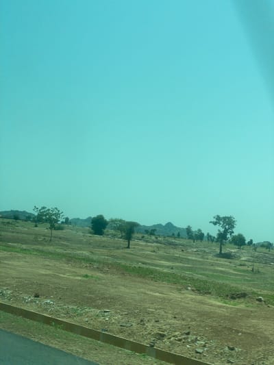 The image captures an outdoor scene with a vast, pale blue sky dominating the left half. The right half of the image displays a dry, grassy terrain with scattered trees. A slightly elevated, dirt path runs diagonally across the land. In the distance, the silhouette of a rocky hill or mountain range is visible against the hazy horizon. The overall impression is of a rural landscape under a bright, possibly hot, sky. The angle of the shot suggests it was taken from a moving vehicle. There is no text visible in the image.