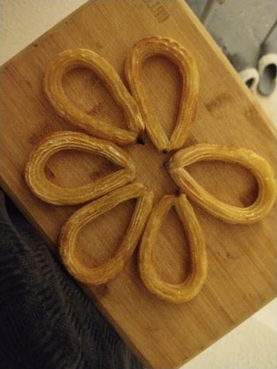 Six golden-brown, teardrop-shaped churros are arranged in a circular pattern on a light brown wooden cutting board. The churros appear slightly glossy, suggesting they may have been fried or coated in a glaze. The board has subtle wood grain visible. In the background, there's a glimpse of a grey slipper and white shoes. A soft, grey fabric is visible on the lower-left of the image, possibly a garment or blanket. The overall composition gives a sense of a simple, homemade treat.