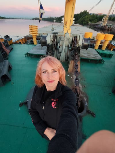 A woman with pink hair takes a selfie on the deck of a ship. She is wearing a black jacket with a pink logo. The deck is green and has various metal structures, including chains and anchor equipment. In the background, a body of water is visible, with a bridge and trees on the far bank. The sky is a soft pink and orange, suggesting either sunrise or sunset. The ship appears to be docked or moving slowly on a river. This scene could be located on the Volga River in Yaroslavl, Russia, as indicated by the presence of industrial infrastructure and the general landscape.
