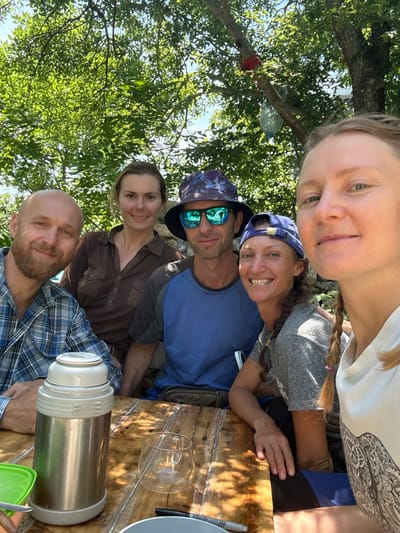 In the city of Chaika, Ukraine, five individuals are captured in a selfie taken outdoors, possibly at a picnic or gathering. The group, consisting of three men and two women, are seated around a rustic wooden table. The man in the foreground on the left has a beard and is wearing a blue plaid shirt. To his right is a woman with brown hair, wearing a brown button-down shirt. In the center, a man in a blue t-shirt and a patterned hat with sunglasses is looking at the camera. Next to him, a woman with a blue cap is smiling. The person taking the selfie, a woman with blonde hair in a braid, is in the foreground on the right. A thermos and a glass are visible on the table, and lush green trees and foliage form a natural backdrop under a bright sky, suggesting a warm, sunny day.