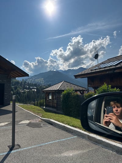 This image depicts a sunny daytime scene in a mountainous residential area of Montriond, France. The foreground shows asphalt pavement with a blue line marking, a black cylindrical bollard, a drainage grate, and a light-colored stone curb bordering a grassy area. On the right, the side mirror of a vehicle reflects a person with light hair wearing a striped shirt, holding a dark smartphone to take the photograph. In the midground, a wooden chalet-style building with a dark roof, satellite dish, and solar panels is visible on the right. To the left, part of a stone building with a wooden roof overhang is present. Centrally, a standalone octagonal wooden gazebo with a dark tiled roof is situated amidst green bushes and grass. The background reveals rolling mountains heavily forested, interspersed with scattered houses, under a bright blue sky containing scattered white cumulus clouds and two visible airplane contrails. The sun is high in the upper left, casting distinct shadows.