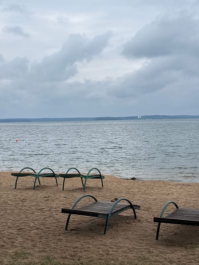 An overcast day is depicted at a beach with several empty lounge chairs on the sand. The water of a large lake stretches to the horizon, with a distant tree line and a few buildings visible on the far shore. A small orange buoy floats in the water. The scene is tranquil and empty, suggesting a quiet time at the beach.