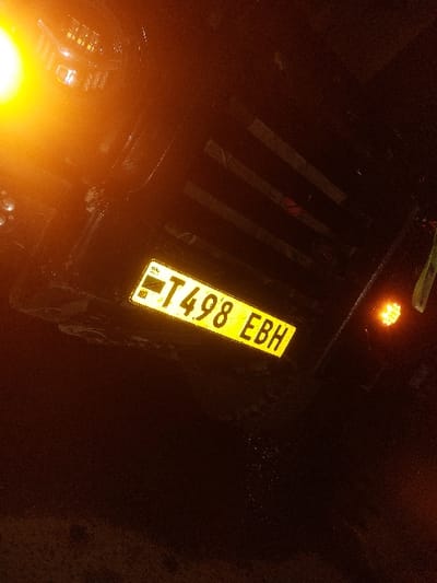 In Msata, Tanzania, a nighttime scene captures the rear section of a dark-colored motor vehicle. The vehicle displays a rectangular yellow license plate with black lettering, oriented diagonally from lower-left to upper-right, reading "T 498 EBH". A prominent, circular orange-yellow light source is visible on the vehicle's left side (image upper-left), with a smaller, amber light on the right side (image mid-right). Multiple horizontal, dark-toned reflective strips are affixed to the vehicle's upper body section, above and to the right of the license plate. The immediate ground below the license plate appears dark and exhibits areas of possible moisture accumulation or reflections. No persons are present; no actions or interactions are discernible.