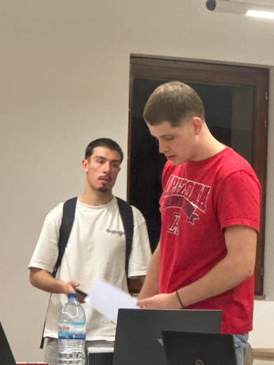 The image depicts an indoor scene with two young adult males in what appears to be an educational or study environment.
In the foreground, on the right side, a male subject with short, dark hair is positioned, looking downwards. He wears a red short-sleeved t-shirt with "ARIZONA" and a letter "A" logo printed in white. A black wristband is visible on his left wrist. His lower body is partially obscured by two dark-colored laptops on a surface; one laptop features a "Dell" logo. He holds several sheets of white paper in his hands.
In the background, on the left side, a second male subject stands. He has dark hair, a mustache, and a goatee. He wears a white short-sleeved t-shirt with faint dark text across the chest (possibly "Samoglou") and a black backpack over both shoulders. He holds a dark-colored smartphone in his right hand and appears to be looking forward.
Between the two individuals, on a surface, a clear plastic water bottle with a blue label (brand "Acova") is visible. The immediate background consists of plain light-colored walls and a dark-framed window or door. Overhead, a horizontal strip light fixture and a small dark circular object are visible on the ceiling. The setting is consistent with a classroom, lecture hall, or study area within a modern educational institution, such as a university campus, located in Guimarães, Portugal. No specific landmarks are visible to identify an exact address or building within the city.