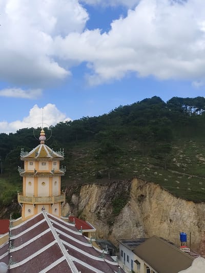 The image shows a view from an elevated position, looking towards a hillside covered in dense green trees under a bright blue sky with scattered white clouds. In the foreground, a multi-tiered, ornate tower, characteristic of Buddhist temple architecture, is visible. The tower is predominantly cream and yellow, with decorative elements and a pointed roof. Adjacent to the tower, a series of reddish-brown tiled roofs with white trim and decorative finials can be seen. Further in the background, a steep, rocky cliff face is partially visible, with some greenery growing on its slopes. At the base of the cliff, a modern building with solar panels on its roof and a blue water tank are present. The overall scene suggests a temple complex situated in a natural, hilly environment, possibly in or near Lộc Quý, Vietnam, given the architectural style and vegetation. There are no people or active interactions depicted in the image.