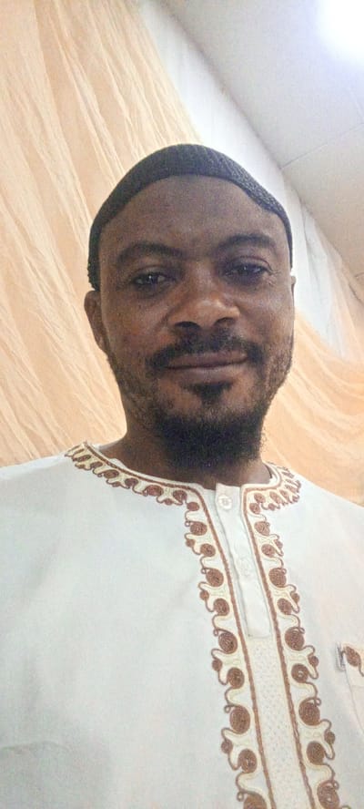 The image is a close-up, waist-up portrait of a man wearing a white thobe with intricate brown and cream embroidery around the neckline and down the front placket. He is also wearing a dark kufi cap. The background consists of beige-colored drapes, which appear to be fabric. The man has dark skin, a short beard, and is looking directly at the camera with a slight smile. The lighting suggests an indoor setting, possibly a home or a place of worship. This image was taken in Gandu Albasa, Nigeria.