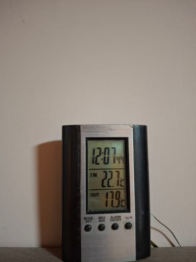 A digital thermometer is placed on a wooden surface against a plain, light-colored wall. The thermometer has a black casing with a silver front panel. The digital display shows the time as "12:07", an indoor temperature of "22.7°C", and an outdoor temperature of "17.9°C". Below the temperature readings are buttons labeled "MODE SET", "MAX MIN", and "ALARM ON/OFF", along with a "°C/°F" indicator. A thin wire extends from the right side of the thermometer, suggesting it has an outdoor probe. The lighting in the scene casts a shadow of the thermometer on the wall. There are no people or animals present. The image is taken from a medium distance, focusing on the thermometer. The background is out of focus. This scene is not specific to Ogre, Latvia; it could be any indoor location with a digital thermometer.