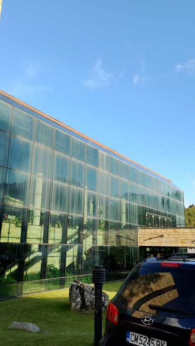 The image depicts the exterior of a modern building with a large glass facade, located in the city of Smolyan, Bulgaria. The building's glass walls reflect the clear blue sky with sparse clouds, as well as the interior of the building, which appears to include plants and architectural elements like stairs and pillars. In the foreground, a black car is parked on a green lawn. A lamppost stands to the right of the car, and several rocks are scattered on the grass. The overall scene suggests a bright, sunny day.