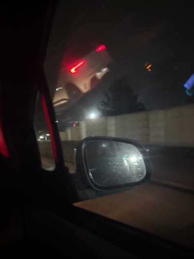 The media file captures a nighttime view from the interior of a vehicle, possibly in Argin, Uzbekistan. The dominant elements are reflections on the car's windshield and the driver's side mirror, suggesting the car is stationary or moving slowly in a built-up area.
Through the windshield, blurred reflections of the car's interior, including a prominent red light strip or trim at the bottom, are visible. Beyond these reflections, indistinct red lights, likely taillights of vehicles ahead, appear in the distance. Other faint, blurred lights and shapes suggest the presence of more vehicles or urban infrastructure.
The driver's side mirror, positioned on the right side of the frame, reflects a brightly lit, obscured scene. The mirror's surface appears somewhat smudged or wet. Within the mirror, blurred green and red lights are visible, along with reflections of what appears to be a multi-story concrete structure, possibly a parking garage, an overpass, or a tunnel wall, with distinct horizontal lines. Bright light sources are also reflected in the mirror.
The overall scene is dark, illuminated primarily by artificial lights and reflections. No specific activity beyond driving or being a passenger in a car at night can be definitively identified. There is no visible text in the image. The setting suggests an urban environment or a road with significant infrastructure.