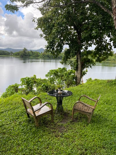 An exterior shot captures a serene lake scene in Si Sunthon, Thailand, under a cloudy sky. In the foreground, two weathered wooden chairs sit on a lush, green lawn. Between them is a small, round, black-topped table with a white vase containing greenery. The chairs face the lake, suggesting a place for relaxation and enjoying the view. The lake itself is calm, reflecting the overcast sky and the surrounding verdant landscape. Trees line the shore, with one large tree with a textured bark positioned prominently to the right of the chairs. In the distance, rolling hills and more foliage complete the tranquil natural setting. The overall impression is one of quiet contemplation and natural beauty.