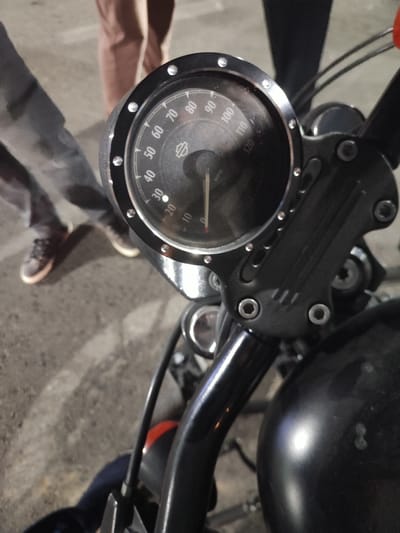 In Dushanbe, Tajikistan, a close-up shot focuses on the dashboard of a black motorcycle. The speedometer, with markings up to 120 MPH, shows the needle just above the 20 MPH mark. A Harley-Davidson logo is visible on the speedometer face. The motorcycle's handlebars and part of its fuel tank are also in view. In the background, out of focus, two individuals are standing, one wearing dark pants and brown shoes, and the other in darker clothing. The ground appears to be asphalt or a similar paved surface. The lighting suggests it is nighttime or a dimly lit area.