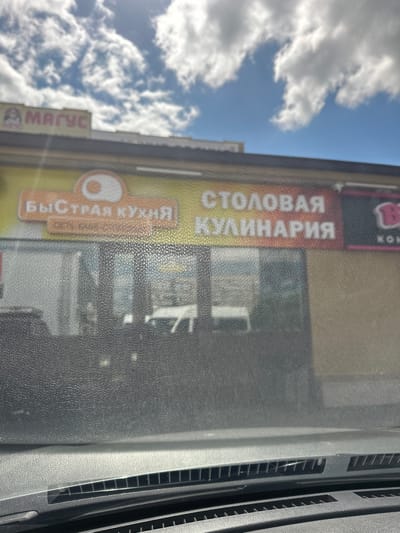 The image shows a storefront with Russian text. The sign above the entrance reads "БЫСТРАЯ КУХНЯ" which translates to "Fast Kitchen" and below that "СТОЛОВАЯ КУЛИНАРИЯ" which translates to "Canteen Delicatessen." To the left of the "Fast Kitchen" sign, another sign reads "МАГУС." To the right of the "Canteen Delicatessen" sign, a portion of a pink sign with a stylized 'B' is visible, possibly indicating a chain like "BystroFood" or similar. The storefront appears to be a commercial establishment in Orsk, Russia. The image is taken from the perspective of someone inside a vehicle, looking out through a dirty windshield. The sky above is blue with white clouds. The focus is on the signage and the storefront.