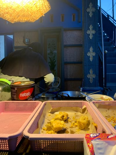 The image depicts a street food stall at night in Tanjungsepreh, Indonesia. A person wearing a dark hijab and apron is preparing food. In the foreground, two pink plastic baskets are visible. The left basket is empty, while the right basket contains fried food items, some of which are on parchment paper. To the left of the baskets, a round tin labeled "nabati Bites" is present, along with a green-lidded jar. In the background, a large woven bamboo light fixture illuminates the scene. A stairway with a dark railing is visible on the right, leading upwards. The overall environment suggests an outdoor food vendor operating after dark.