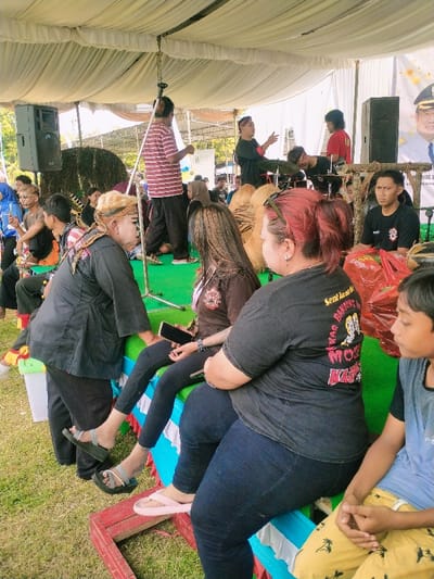 The scene is a gathering of people under a large white tent. The tent is supported by metal poles. There are multiple people, seated or standing, with the majority of the people seated on benches with a blue and white striped covering. There are a few men standing at the back of the tent, near a large speaker. The people are situated on green grass. There is a wooden platform with a red and white striped covering near the front of the scene. The coordinates (latitude: -7.47, longitude: 111.064) place the location in a suburb of Karangmalang, Indonesia.