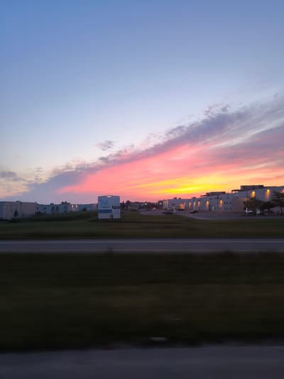 An exterior, full shot captures a sunset over a commercial area in Mirabel, Canada. The sky transitions from a soft blue at the top to vibrant hues of pink, orange, and yellow near the horizon, with wispy clouds scattered throughout. In the mid-ground, several low-rise industrial or commercial buildings with flat roofs line the landscape. One prominent building on the right features numerous evenly spaced lights illuminating its exterior walls. To the left of this building, a large white sign with a blue "M" logo is visible. A strip of green grass separates the buildings from a highway in the foreground, with a blurred motion effect suggesting the photo was taken from a moving vehicle. The highway itself is dark asphalt with white lane markings. The overall scene depicts a serene evening in an urban industrial setting.