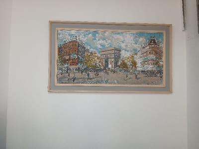 A framed painting depicting a cityscape hangs on a white wall. The painting portrays a bustling city square featuring the Arc de Triomphe in the background. Buildings with intricate architecture flank the square, and numerous small figures represent people moving about. The sky is filled with clouds, and the painting is framed with a light gray wooden frame.