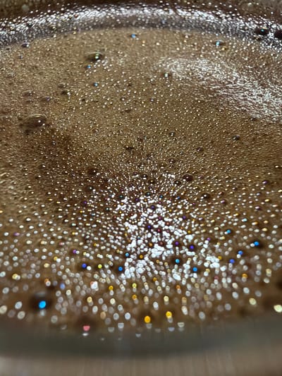 The image is a close-up, macro shot of a surface covered in small, shimmering bubbles. The bubbles are predominantly brown, suggesting a liquid like coffee or chocolate, with some exhibiting iridescent highlights in shades of blue, purple, yellow, and white due to light reflection. The texture is foamy and appears wet. The perspective is from above, looking down at an angle onto the bubbly surface. The lighting creates a sparkling effect on the bubbles, with some areas appearing brighter and more out of focus than others, indicating a shallow depth of field. The overall impression is one of abstract beauty and texture. There are no people, explicit objects, or discernible scene or setting beyond the bubbly surface itself. The image does not convey any specific time of day, weather, location cues, or emotions. There is no visible text in the image. The context of "Simonikha, Russia" is not visually represented in the photograph.