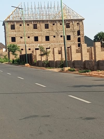 The image shows a multi-story building under construction on a street in Garko, Nigeria. The building is made of brick and appears to be several floors high, with exposed rebar on the roof, indicating it is not yet completed. A dark-colored roof is visible on a lower structure adjacent to the main building. The street in the foreground is paved asphalt with white lane markings. Several streetlights are present, with one prominently positioned in front of the construction. Trees and vegetation are scattered around the building site, and some construction materials, possibly bricks or concrete blocks, are visible along the perimeter of the property. The sky is clear and blue, suggesting fair weather. There are no people or vehicles visible in the image. The scene is an urban environment with ongoing construction work.