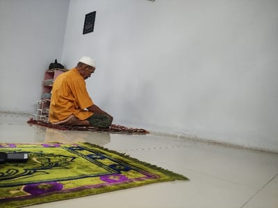 An indoor scene captures an adult man engaged in Islamic prayer, kneeling on a prayer mat in a simple room. The man, with grey or white facial hair, wears a white skullcap (kufi) and an orange-yellow long-sleeved shirt over green or patterned lower garments, possibly a sarong. He is positioned facing right in the image, with his body in a focused, reverent posture typical of *salat*, likely *jalsa* (sitting between prostrations) or a similar kneeling position.

He is situated on a small, reddish-brown patterned prayer mat, which is placed atop a larger, vibrant green prayer mat featuring intricate purple and dark blue floral or geometric patterns. To his right, on the green mat, a small dark rectangular object (possibly an electronic device or speaker) is visible. Behind him, a white and green striped woven bag or basket is partially obscured.

The setting is a plain room with white walls, creating a minimalist backdrop for the spiritual activity. A corner where two walls meet is clearly visible, and a small, dark-framed object, possibly a picture or religious scripture, hangs on the wall in the lower left of the frame. The lighting appears soft and ambient, suggesting an indoor environment during an unspecified time of day. The scene evokes a sense of peace and devotion, consistent with prayer in a private or semi-private setting in Situbondo, Indonesia. No discernible text is visible on the framed object due to distance and resolution.