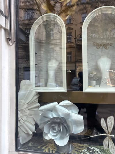 A window display features two framed, arched cutouts, each showcasing a white vase. Reflections of buildings and trees are visible on the glass surfaces. To the right, a large, stylized white paper rose and a fan-like paper flower accent the display. A white dragonfly sits above the rose. The scene is viewed from a slightly upward angle.