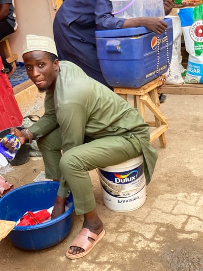 This image captures a vibrant market scene in Kafanchan B, Nigeria, showing a man engaged in an activity within an informal outdoor setting. The photograph is oriented sideways, giving an overhead perspective of the subjects and their surroundings.
The central subject is a man, likely in his 20s or 30s, dressed in a light green tunic and trousers, and wearing a white kufi cap. He is crouched down, looking directly at the camera with a neutral to slightly curious expression. His hands are positioned over a blue plastic basin, where a red object, possibly a tool or clamp, is visible amidst some liquid or suds. He holds a small blue and yellow container, partially identifiable by "Tide" branding, in his left hand. His right foot, wearing a light brown sandal, is visible next to the basin.
Beside him, a white Dulux paint bucket is prominent, bearing the text "Dulux Silk Emulsion Walls & Ceilings AkzoNobel." In the background, to the left, another person's arms in dark blue attire are visible, holding a large blue cooler adorned with a "Pepsi" logo and a string of blue and white beads, possibly a rosary. The background features various large sacks of goods, with visible text including "GOLDENA FOO," "The NOW GOOD E," and "DEN PENNY NGAR Ane Fortified." Wooden structures, possibly makeshift market stalls or furniture, are also scattered around. The ground is a mix of dirt and concrete, showing some damp patches. The scene suggests a busy marketplace or an informal trading area during daytime.