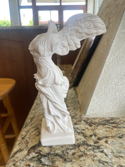 A white, classical-style statue depicting a winged figure, reminiscent of the Winged Victory of Samothrace, is prominently displayed indoors in Markovo, Bulgaria. The statue, likely made of marble or a white composite material, is oriented horizontally due to the image's rotation, with its base resting on a mottled beige and brown granite surface.

The statue features finely detailed wings with sculpted feathers and flowing drapery around the figure's body, emphasizing movement and grace. Its head is turned towards the left, with an arm (partially obscured or missing, consistent with ancient sculptures) extending forward. The base of the statue is a simple rectangular block.

Behind the statue is a reflective surface, possibly a mirror or polished glass, which reflects parts of the room, including window frames and bright outdoor light. Above this reflective surface is a light-colored, textured wall or ceiling. To the left of the statue, through visible windows, a bright outdoor scene with soft green foliage is discernable, indicating it is daytime. Further back in the room, darker wooden elements, possibly part of a door or furniture, are visible. The granite surface on which the statue sits exhibits a natural pattern of varying shades of brown, beige, and black. No visible text is present in the image. The scene suggests a decorative display within a home or public building.