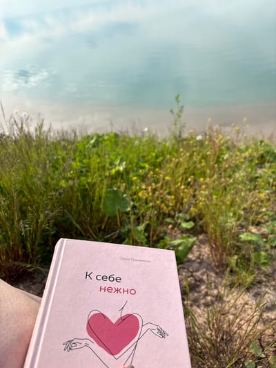 The image depicts an outdoor scene located in Sapunov Bor, Russia.

In the background, a serene body of water, likely a lake or large pond, exhibits a light blue-green hue. The water's surface reflects the sky, showing diffuse white cloud formations against a pale blue expanse, indicative of an overcast or soft light condition. A narrow strip of light-colored, possibly sandy, shoreline is visible where the water meets the land.

The midground features dense, wild vegetation, including tall, thin green grasses on the left and various broad-leafed plants with small yellow flowers scattered towards the right. The ground beneath the foliage appears dry and somewhat cracked.

In the foreground, the upper portion of a human leg or thigh is visible on the lower left, appearing light-skinned. The person is holding a light pink book prominently displayed and in sharp focus. The book's cover features Russian text: "Ольга Примаченко" (Olga Primachenko) as the author, and "К себе нежно" (To self gently) as the title, with "нежно" highlighted in red. An abstract line drawing on the cover depicts the outline of a female torso from the chest to the waist, encompassing a large, solid red heart shape. The hands of the figure are subtly outlined below the heart. The scene suggests a quiet moment of reading and relaxation in a natural, serene environment.