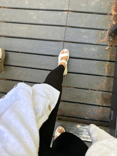A first-person perspective shot shows a person's legs and feet on a wooden deck. The person is wearing black leggings and white sandals. A white textured shirt is draped over their left leg. The deck is made of weathered gray wooden planks, and fallen pine needles are scattered across it. The environment appears to be outdoors, possibly in a park or natural area in Morskoe, Russia, given the presence of pine needles and the rustic wooden structure. The lighting suggests it is daytime.