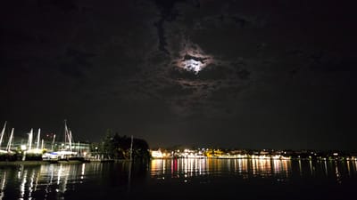This is a nighttime photograph taken from the water, looking towards a marina and a town. The sky is dark with dramatic clouds, partially obscuring a bright full moon that casts an ethereal glow. The water in the foreground is calm, reflecting the lights from the shore and the moon.

On the left side of the frame, a marina is visible with numerous sailboats docked. Their tall masts and rigging are silhouetted against the illuminated buildings behind them. Lights from the buildings and from the boats reflect in the water, creating shimmering patterns of white, yellow, and red.

To the right, the shoreline features a town or city with a dense collection of buildings. These buildings are illuminated, outlining the cityscape against the dark sky. The reflections of the town's lights are prominent in the water, creating a colorful and dynamic effect. Trees are also visible along the shoreline, adding a touch of greenery to the scene.

The overall atmosphere is serene and magical, with the moonlight and the reflected lights creating a sense of enchantment. The time of day is clearly night, and the presence of the moon suggests a clear night, although the clouds add a dramatic element. The location cues, such as the sailboats and the Mediterranean-style architecture, suggest a coastal or lakeside setting, consistent with Peschiera del Garda, Italy. No people are explicitly visible, focusing the viewer's attention on the interplay of light and water.