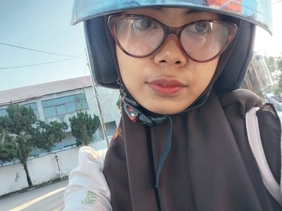 This is a selfie taken by a young woman wearing a blue helmet and brown hijab, with large, reddish-brown glasses. She is looking at the camera, and her lips are slightly parted, with a hint of red lipstick. The woman is on a motorcycle, as evidenced by the helmet and the blurred background of trees and a building. The building appears to be a factory or warehouse with a light-colored wall and a tiled roof. There are power lines visible against the light blue sky, suggesting it is daytime. The overall mood is casual and perhaps a bit candid, as she is looking directly into the camera. The scene is outdoors, with sunlight casting shadows on the ground, indicating it's likely a sunny day. The location is identified as Macege, Indonesia.