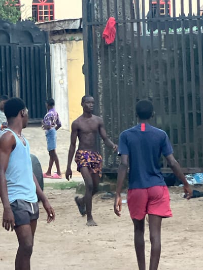 The scene is outdoors in Ijegun, Nigeria. Four Black males are visible, seemingly engaged in physical activity. The first, wearing a blue shirt and red shorts, is at the top of the image. The second male is shirtless, wearing patterned shorts. The third male wears a patterned shirt and blue shorts with pink sandals. The fourth male wears a blue tank top and dark shorts, positioned near a tire. A metal gate is in the background, along with a building with red window frames. A red cloth hangs from the gate. The ground is sandy or dirt-covered.