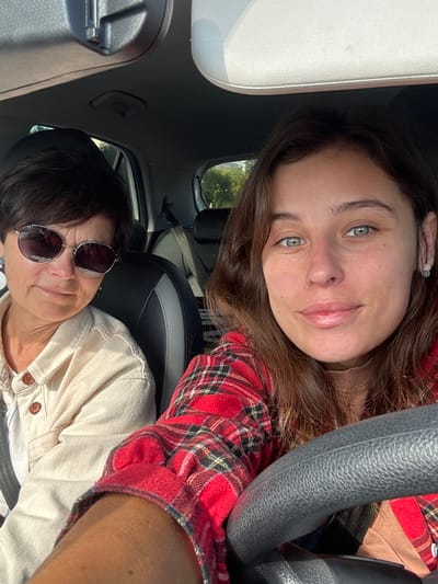 The image is a selfie taken inside a car. Two women are visible. The woman in the foreground, on the right, has brown hair and is wearing a red and black plaid shirt. She is looking at the camera and smiling. The woman in the background, on the left, has short dark hair and is wearing sunglasses and a light-colored shirt. She is also looking at the camera. The car's steering wheel is visible in the foreground. The interior of the car appears to be modern. The lighting suggests it is daytime, with sunlight coming in from outside. The background visible through the car windows shows trees and greenery, indicating an outdoor environment. The image does not contain any specific landmarks or elements that would allow for the precise identification of its location within Jeju City, South Korea, other than it being a car.
