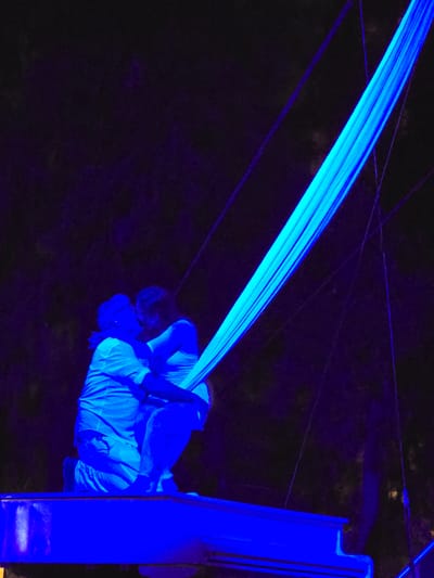 In Cornellà de Llobregat, Spain, a romantic aerial performance unfolds under a deep blue light. Two individuals, one kneeling and embracing another who is seated, are suspended by a bright blue fabric that hangs diagonally across the frame. They appear to be kissing, their forms silhouetted against the dark, possibly outdoor, background. Below them rests a portion of a white grand piano, illuminated by the same intense blue light. The scene suggests a theatrical or artistic presentation, with the surrounding darkness and the suspended fabric creating a dramatic and intimate atmosphere.