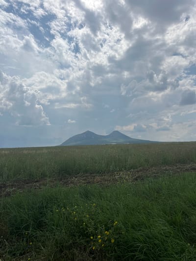 The image shows a landscape view of two peaks in the distance, likely mountains, under a cloudy sky. In the foreground, there is a field of green grass with some yellow wildflowers and patches of dark soil. The sky is partly cloudy with dramatic formations of white and grey clouds, suggesting fair weather with potential for rain. The overall scene depicts a rural or natural environment. The location is specified as Dunayevka, Russia.