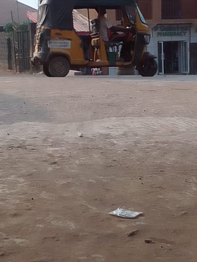 A daytime forensic image captures a street scene in Nkwelle Ezunaka, Nigeria. Centrally positioned on an unpaved, light brown dirt road is a yellow motorized tricycle, commonly known as a Keke Napep, fitted with a dark green canopy. One individual, dressed in a white short-sleeved top and dark trousers, is seated in the driver's position, hands on the handlebars. The tricycle exhibits a weathered appearance on its rear section, with a white sticker containing text affixed to the rear left. A green container is visible within the passenger area.
In the background, to the right of the tricycle, a light-colored multi-story building features a sign clearly identifying "Goldfox PHARMACY" above its glass entrance doors. To the left background, a partial view of a concrete block wall and a metal gate with a reddish-brown roof structure behind it is discernible. The foreground consists of dry, uneven earth with scattered small stones and patches of lighter dust. A crumpled, light-colored wrapper, approximately rectangular in shape, lies on the ground in the immediate foreground, slightly to the right of the center. A small red object rests on the ground behind the tricycle. The scene is illuminated by diffuse daylight.