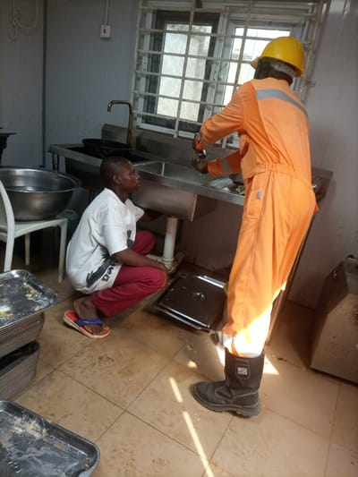 An indoor scene in Opete, Nigeria, depicts two individuals in what appears to be a utility or kitchen area. The environment features light gray walls, a tiled floor, and a window with metal security bars. A large, double-basin stainless steel sink unit with a gooseneck faucet is centrally positioned.
On the right, an adult male, wearing an orange full-body coverall with reflective stripes on the back and a yellow hard hat, stands facing the sink. He is operating the faucet or engaged in washing an item in the right sink basin. He wears black work boots.
To the left, a younger individual with dark skin and short hair crouches on the floor, wearing a white short-sleeved t-shirt with a dark graphic, red pants, and red and blue flip-flops. This individual is looking towards the adult male and the sink area.
Adjacent to the crouching individual, a white plastic chair holds a large metal bowl filled with water. Several stacked rectangular metal trays, some containing food residue, are visible on the floor in the foreground. Sunlight enters through the window, casting highlights and shadows across the floor and objects.