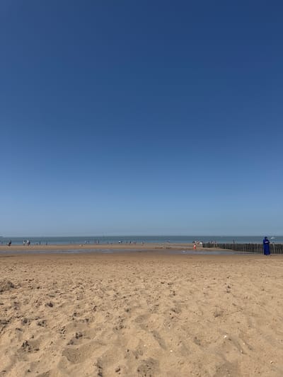 A photograph shows a beach scene in Knokke-Heist, Belgium. The sky takes up half of the shot and is clear blue. The right half consists of a sandy beach that merges into the sea. People are seen along the water's edge, some appearing as blurred figures. Wooden breakwaters are present along the coastline.