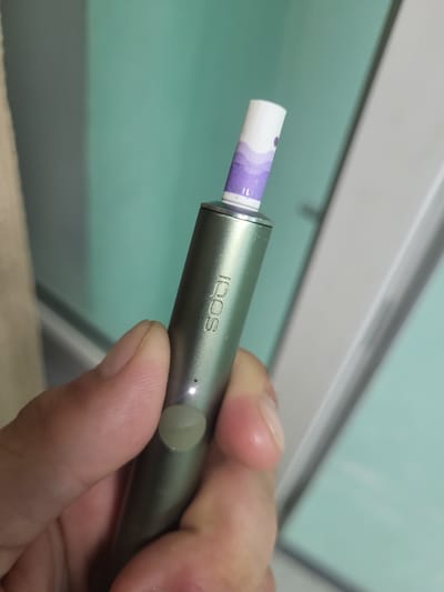 A close-up shot shows a person's hand holding a metallic green vaping device. The device has the brand name "IQOS" inscribed on its side. A light is visible on the device, and a button is present near the bottom. At the top of the device, a white and purple tobacco stick is inserted. The background consists of a wall with a gradient from dark gray to teal, and a white vertical structure. The image was taken in Khotsky, Ukraine.
