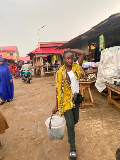 The image captures a scene in Kafanchan B, Nigeria. A woman stands in the foreground wearing a yellow patterned shirt, a white top, and checkered pants. She has a black purse slung across her shoulder and is holding a clear plastic container. The background reveals a bustling marketplace with stalls and people. There is a building with a red roof and "POS" written on it. A person on a motorcycle and a woman in a blue robe are also visible in the lower part of the frame. The ground appears to be paved or made of concrete. The weather seems overcast.