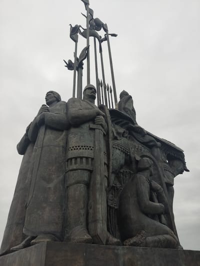 This is a low-angle, medium shot of a bronze monument located in Khotitsi, Russia. The sky is overcast. The monument features multiple human figures, mostly warriors dressed in armor and robes. Many of the figures are holding long spears with banners or pendants attached near the tips. One warrior appears to be on horseback at the top of the monument. The sculpture depicts a scene of warriors in a historical or mythical context.