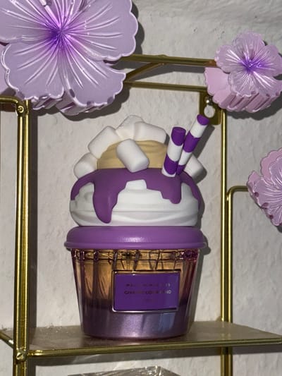 This image, located in Vellmar, Germany, depicts a decorative object resembling a dessert on a golden metallic shelf, against a textured, light-colored wall. The primary subject is a purple and white dessert-themed ornament, appearing to be a sundae or milkshake. It features a beige top layer adorned with multiple white cylindrical marshmallow-like pieces and two purple and white striped stick ornaments. Below this is a white rounded section with purple syrup-like drips. The base is a reflective, ribbed purple container, possibly plastic or glass, shaped like a cupcake or sundae glass. A rectangular label on the container's front displays white text on a purple background, partially legible as "MALLOW MADNESS" and "GIVE ME GOODNESS". Surrounding the main object and attached to the golden stand are several translucent, light purple, flower-shaped decorations. No people, actions, or interactions are visible.