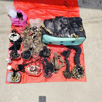 An outdoor scene captures a collection of items laid out on a vibrant red tarp, which covers about two-thirds of a light gray concrete surface. The strong sunlight casts distinct shadows, indicating a daytime setting with clear weather.

Various objects, likely components for traditional crafts, jewelry, or spiritual items, are displayed. A prominent black and light blue duffel-style bag occupies the center-right, adorned with gold-colored bells and white cowrie shells attached to black cords. Partial text on the bag reads "BIAS..." and "ENTERPRISE."

Numerous bundles and coils of black cord and thread are arranged across the tarp. Some cords are decorated with white rectangular pieces, possibly bone or shell, while others feature clusters of small, dark reddish-brown seeds (resembling Rudraksha beads) or light-colored cylindrical beads. Gold-colored bells are also seen in a small gray plastic tray, attached to black threads.

Scattered among these are substantial quantities of dried plant matter, roots, and small seeds, suggesting natural elements used in crafts or traditional practices. A pair of scissors is partially visible amidst these dried materials. Several small plastic containers, including red rectangular trays and a brown bowl, hold coiled black cords, dark elongated pieces, and more decorative elements. Two small white plastic bottles are also present on the left side of the tarp.

In the top left, a pink fabric bag holds additional black cords and a clear plastic bag. Larger, weathered pieces of wood or roots are also distributed among the items. The overall arrangement suggests a market stall or a preparation for selling goods, consistent with the cultural context of Lalmohan, Bangladesh.
