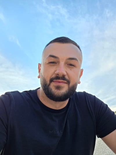 A close-up, low-angle, outdoor selfie shows a man with short, dark hair, a neatly trimmed beard and mustache, and light-colored eyes. He is wearing a black t-shirt with a subtle dark logo on the left chest. The man is looking directly at the camera with a slight smile. The background is a pale blue sky with wispy white clouds. The bottom edge of the image shows a rough, speckled surface that appears to be gravel or sand. The image is taken in the city of Suvorovo, Bulgaria.