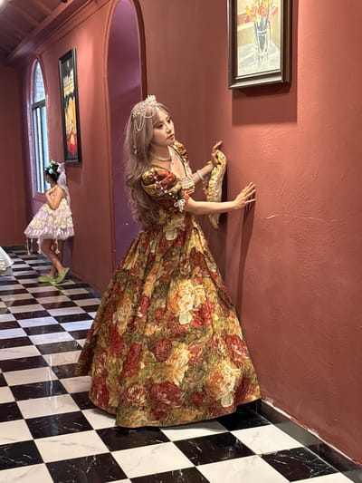 A woman in a floral gown and crown is standing in a room with black and white checkered flooring. A painting of flowers hangs on a red wall. Another painting of a woman is visible in the background, near a window. There is a woman in a light purple dress behind the main figure. The location is Baqiao, China.