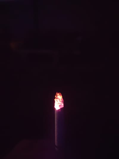 A close-up, dark image shows the glowing ember of a cigarette in the bottom center. The ember is a vibrant red-orange and casts a subtle pinkish light on the surrounding cigarette tip. The rest of the image is black, with a slight gradient suggesting a dimly lit or dark environment. There is no discernible location in Northampton, United Kingdom.
